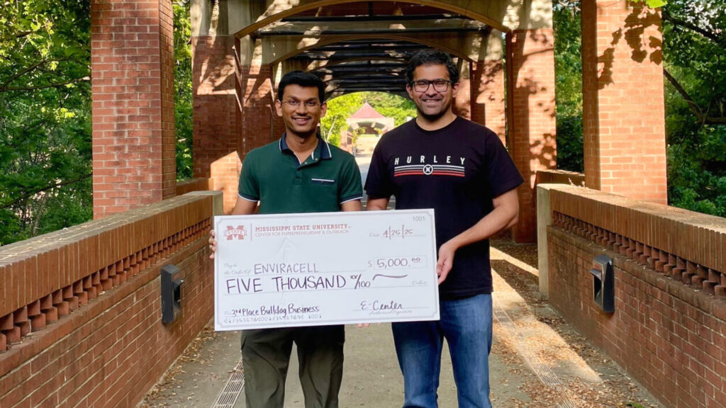 Two people standing on a brick walkway holding a large check.